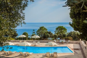 Una piscina con sillones de descanso y sombrillas, rodeada de árboles y vistas al mar.