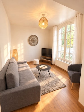 Ein Wohnzimmer mit einer Couch, einem Fernseher, einem Teppich und einem Fenster mit Vorhängen.