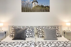 A double bed with patterned bedding and heart-shaped pillows, a nightstand with a lamp, and a wall-mounted picture of a lighthouse. - Holiday apartment with 1 bedroom (Timmendorfer Strand)