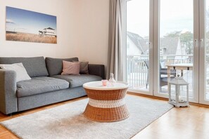 A living room with a grey sofa, a round wicker coffee table, a white rug, and a view through a glass door. - Holiday apartment with 1 bedroom (Timmendorfer Strand)
