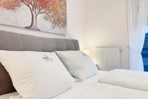 A hotel room with a bed, pillows, a radiator, and a window with curtains. - Holiday apartment with 1 bedroom (Timmendorfer Strand)