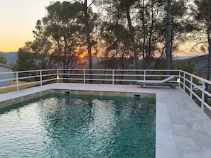 A swimming pool with a white deck and a white railing, surrounded by trees and a sunset in the background. - Villa Bohemia: Sea View, Private Pool, and Wi-Fi (Olivella)
