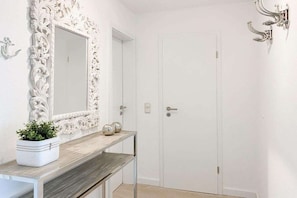 A hallway with a white door, a mirror with an ornate frame, a small table with a potted plant, and a wall-mounted light fixture. - Holiday apartment with 2 bedrooms (Timmendorfer Strand)