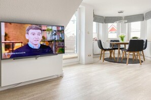 A modern living room with a flat-screen TV mounted on a white cabinet, a dining area with a round table and chairs, and a large window providing natural light. - Holiday apartment with 2 bedrooms (Timmendorfer Strand)