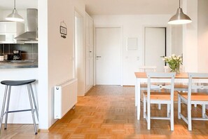 A modern kitchen with a white countertop, a black bar stool, and a dining area with a wooden table and white chairs. - Holiday apartment with 1 bedroom (Timmendorfer Strand)
