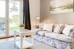 A living room with a sofa, a coffee table, a lamp, and a painting on the wall. - Holiday apartment with 1 bedroom (Timmendorfer Strand)