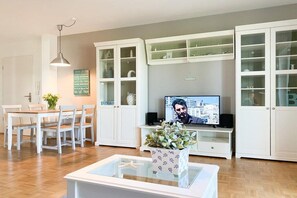 Un saló amb televisió, armaris blancs, una taula de dinar amb cadires i una taula de centre de vidre amb una planta.