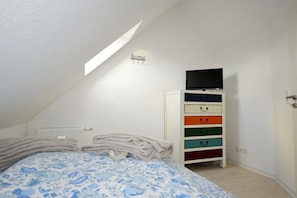 Une chambre avec un lit, une armoire blanche dotée de tiroirs colorés, un téléviseur posé sur le dessus, et un panneau indiquant « chez soi » accroché au mur.
