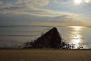 Ein Strand mit einer langen, schmalen Mole, die ins Wasser führt, einem klaren Himmel und einer tief stehenden Sonne am Horizont.
