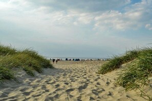 Ein Sandweg am Strand, gesäumt von Dünen, der zu einem Strandbereich mit Personen und Strandausrüstung führt.