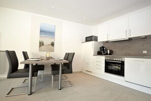 A modern kitchen with white cabinets, a dining table with chairs, and a view of the beach through a large window. - Holiday apartment with 1 bedroom (Norddeich)