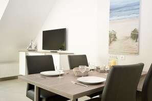 Ein Essbereich mit Tisch für vier Personen, einem an der Wand befestigten Flachbildfernseher und einem Strandbild über dem Tisch.