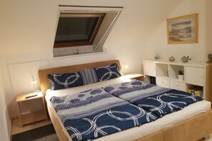 A bedroom with a bed, a nightstand, a bedside table, a shelf with decorative items, and a window with a view. - near Warnemünde (Rostock) (Admannshagen-Ausbau)