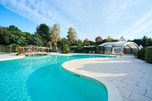 A swimming pool with a curved edge, surrounded by lounge chairs and umbrellas, with a covered area and trees in the background.