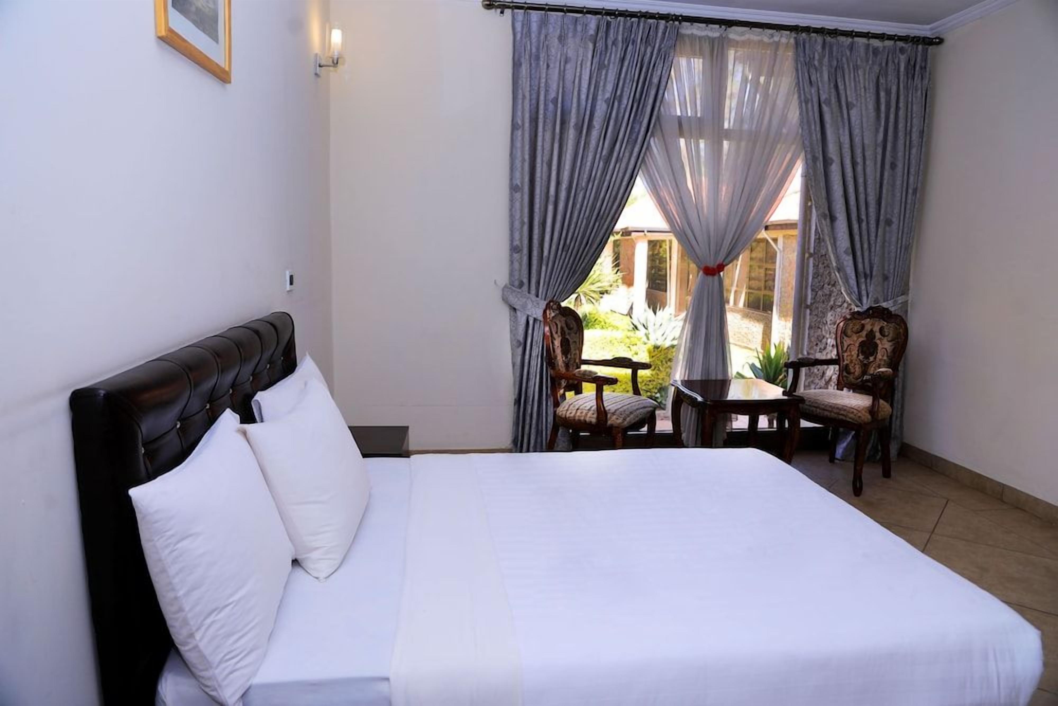 A hotel room with a bed, a table, two chairs, and a view of a garden through sheer curtains.