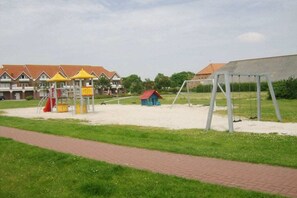 Une aire de jeux avec un toboggan et des balançoires, entourée d’un chemin en briques et de verdure.