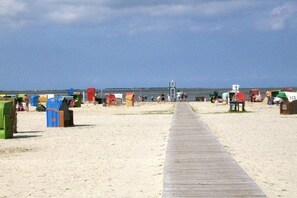 Une plage avec une promenade en bois menant à la mer, entourée de cabanes de plage et sous un ciel dégagé.