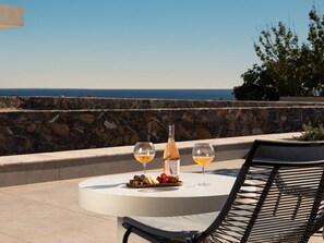 A round table with a bottle and two glasses of wine, a plate with food, and a chair, overlooking a stone wall and the sea. - Villa in Ierapetra With Swimming Pool (Ierapetra)