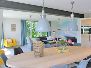 Ein moderner Essbereich mit einem großen Holztisch, mehreren Stühlen, hängenden Pendelleuchten und Blick ins Freie durch große Fenster.