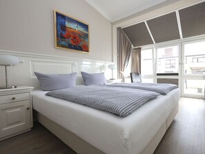 A hotel room with a large bed, a bedside table with a lamp, a painting on the wall, and a view through a window. - 28 Residence Bismarck (Sylt)