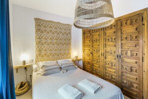 Un dormitorio con cama, lámparas de noche, cabecera tejida, armario de madera y una lámpara colgante de fibras naturales.