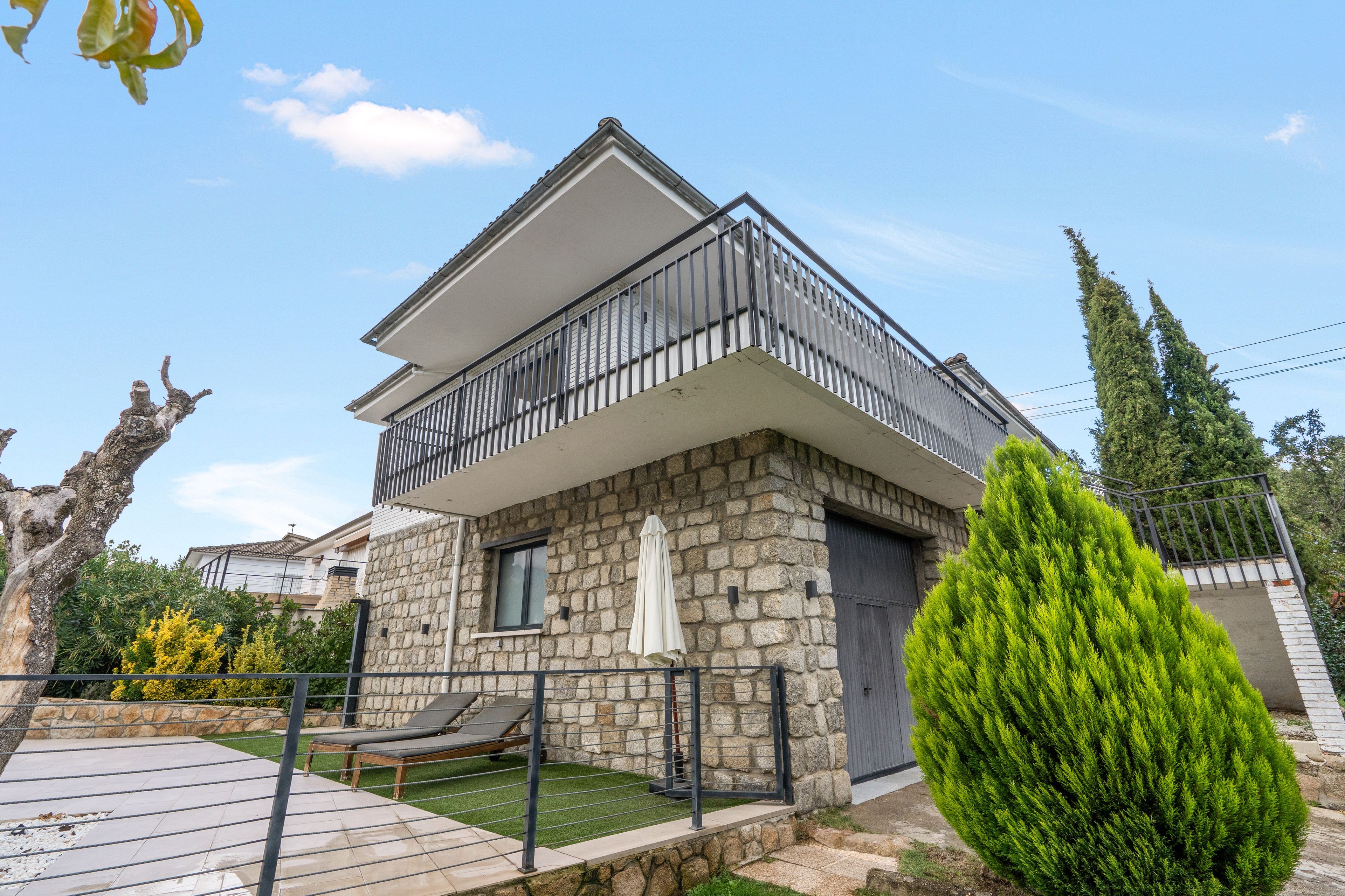 A two-story house with a balcony, stone facade, and a small patio with a lounge chair.