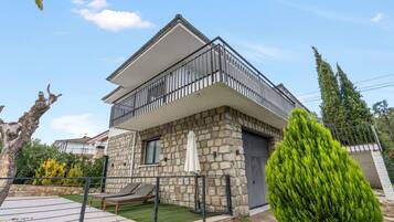 A two-story house with a balcony, stone facade, and a small patio with a lounge chair.