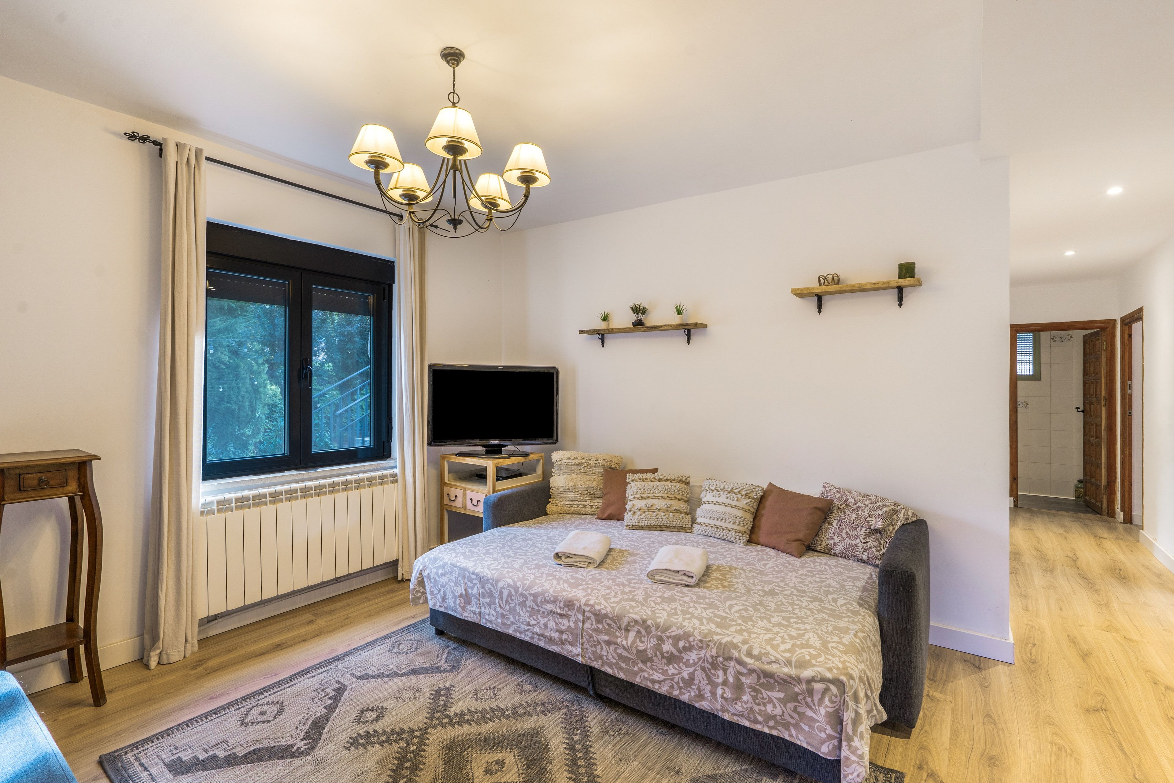 Een slaapkamer met een groot bed, een televisie, een raam met gordijnen, een kroonluchter en een houten vloer.