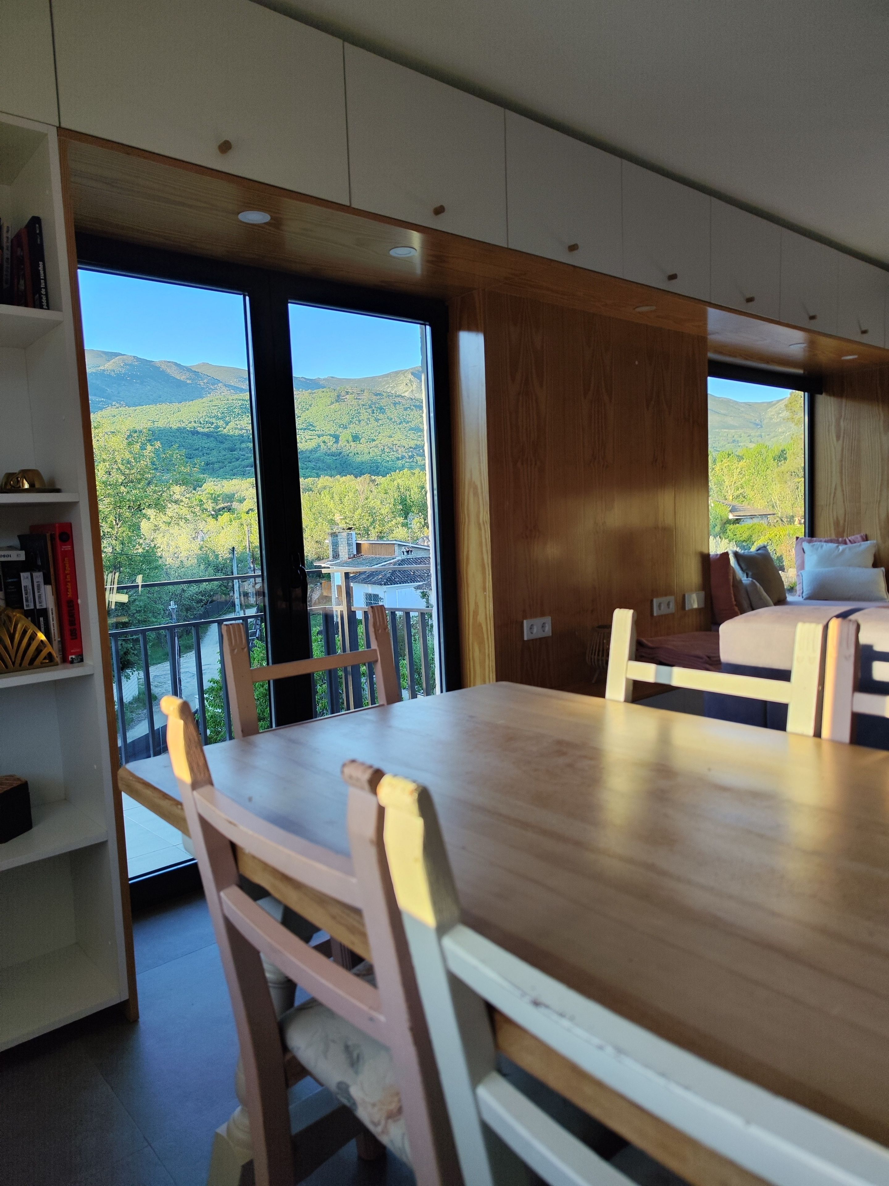 Un interior moderno con una mesa de comedor y sillas, una estantería y ventanas amplias con vistas a un paisaje montañoso.