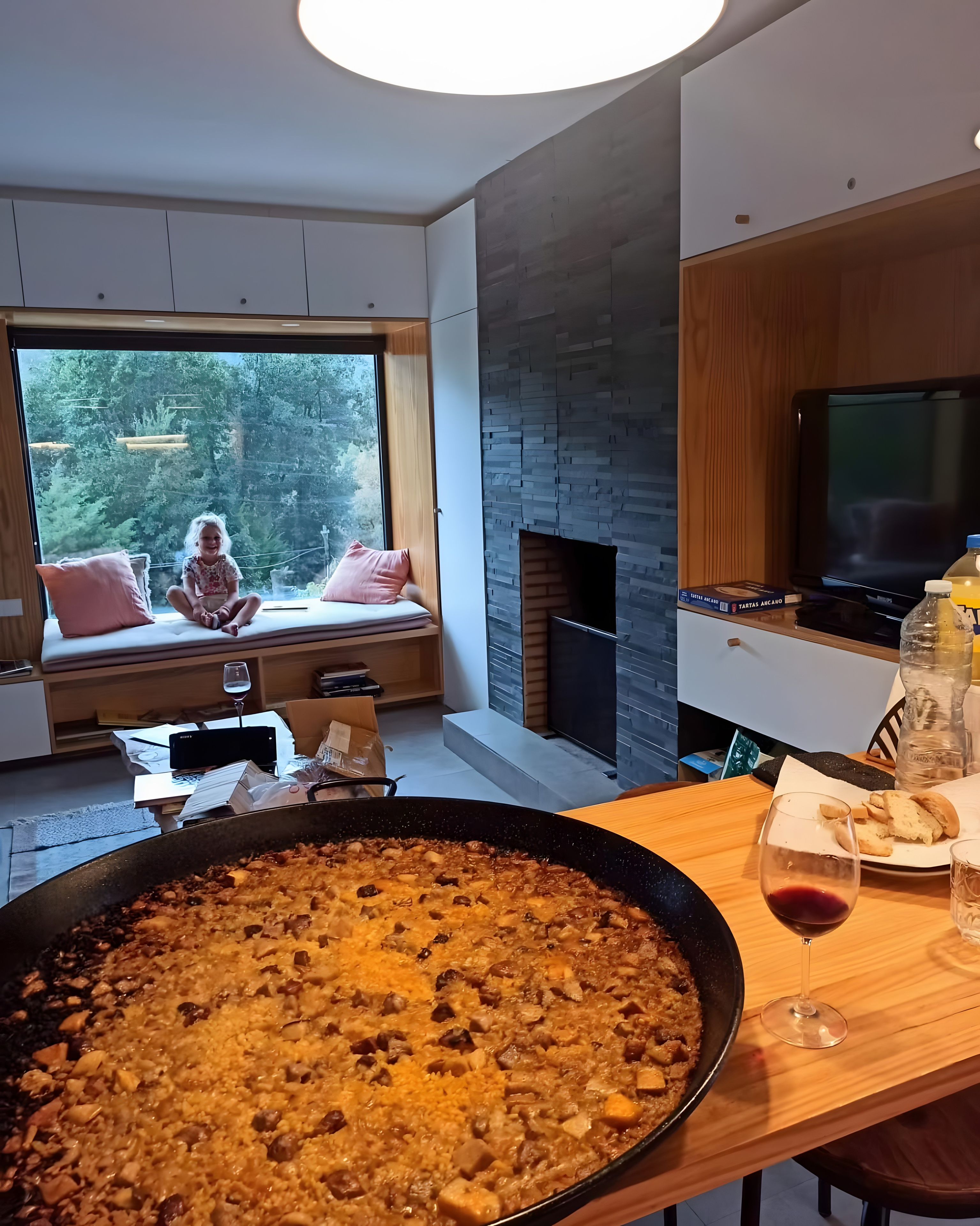 Ett modernt vardagsrum med ett stort fönster, en öppen spis och en matplats där en stekpanna med mat står på spisen.