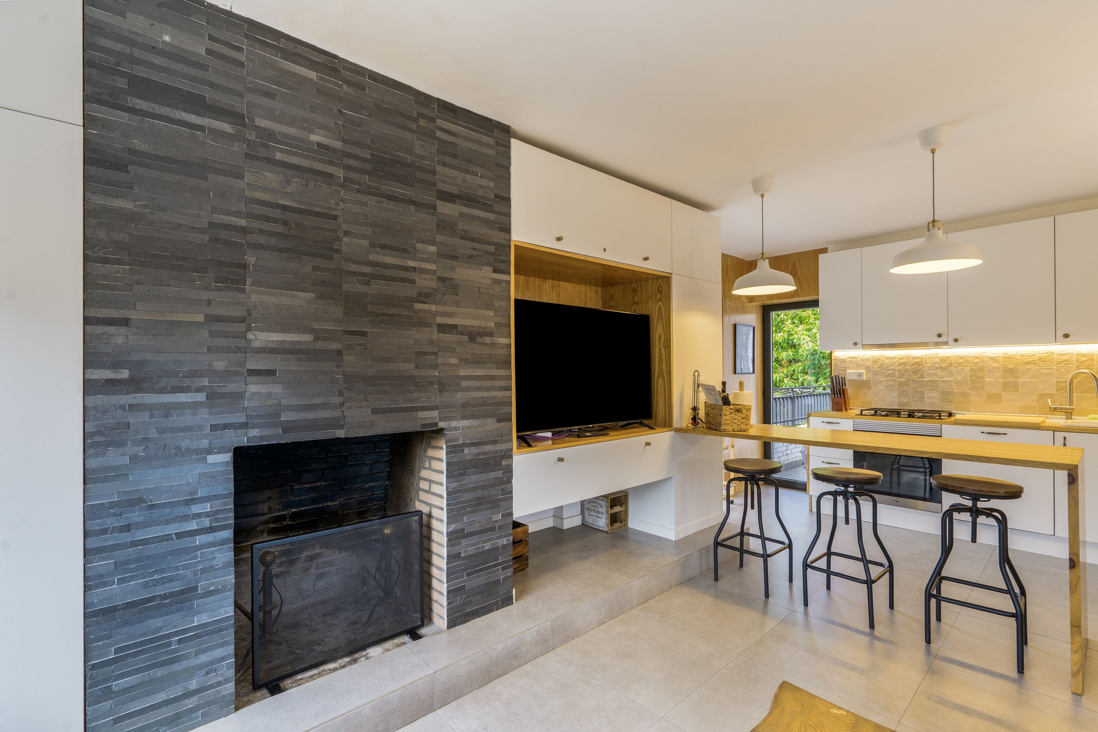 A modern kitchen with a stone fireplace, a large window, and bar stools.