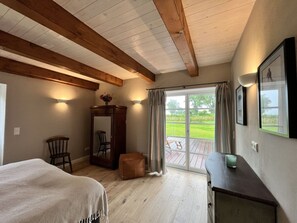 Ein Zimmer mit einem Bett, einem Holzstuhl, einem kleinen Tisch mit einer Vase und Blick auf eine Terrasse durch eine Glastür.