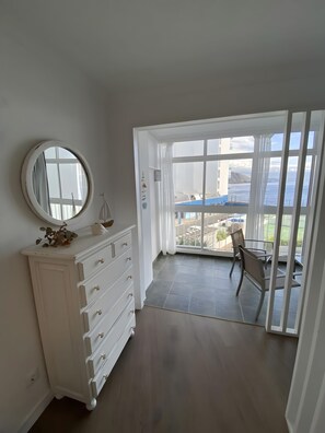 Ein Zimmer mit einem weißen Schminktisch, einem runden Spiegel und Meerblick durch große Fenster.