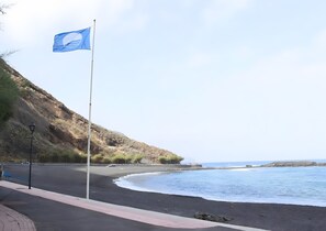 Eine Küstenstraße mit Fahnenmast, einem schwarzem Sandstrand und einem felsigen Hügelhang.