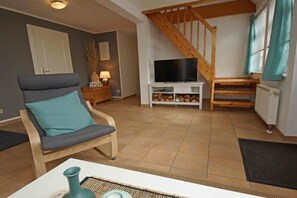 Una sala de estar con una silla de madera, un televisor sobre un mueble, una escalera y una puerta.