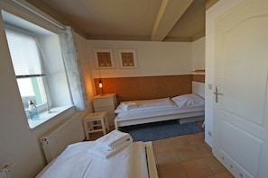 Una habitación pequeña y limpia con dos camas, una ventana con cortinas, un radiador y una lámpara fijada a la pared.