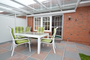 Eine Terrasse mit Glasdach, einem weißen Tisch und grün gestreiften Stühlen.