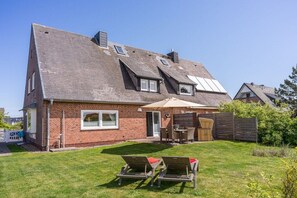 A two-story house with a brick exterior, a sloped roof, and a small backyard with a patio and two lounge chairs. - Holiday home with 1 bedroom (Wenningstedt)