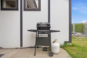 Una parrilla portátil y un tanque de propano frente a un edificio con fachada blanca y detalles negros.