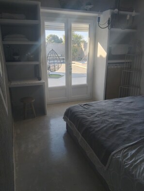 Ein Schlafzimmer mit einem Bett, einem kleinen Hocker, einer Regaleinheit und einer Glastür, die auf einen Balkon führt.