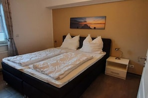 Ein ordentlich bezogenes Bett mit weißen Leinen und Kissen, ein Nachttisch mit Lampe sowie ein Sonnenuntergangsgemälde an der Wand.