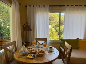 Una tavola imbandita per la colazione con diversi cibi e bevande, una sedia, una lampada e vista sul verde attraverso la finestra.