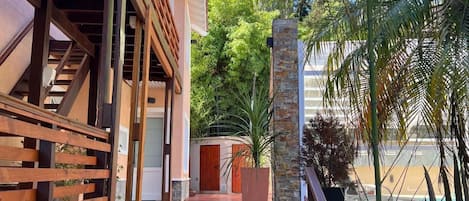 A wooden staircase leading to a covered outdoor area with a stone pillar, surrounded by greenery and a clear sky.