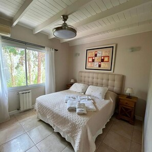 A bedroom with a bed, a ceiling fan, a radiator, a window with curtains, a bedside table with a lamp, and a framed picture on the wall. - Alojamiento Carilo Princes (Carilo)