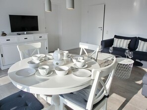 Ein modernes Wohnzimmer mit einem runden Esstisch, der für Tee gedeckt ist, einer Couch und einem Fernseher im Hintergrund.