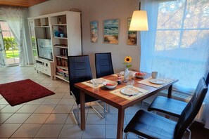 A dining area with a wooden table, black chairs, and a red rug. There are paintings on the wall, a window with a view, and a white cabinet with shelves. - Holiday home in Hooksiel with swimming pool (Hooksiel)