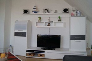 A living room with a flat-screen TV, white entertainment unit, and shelves with decorative items. - Holiday apartment with 2 bedrooms (Hooksiel \/ Wangerland)