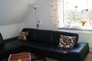 Un sofà de pell negra amb dos coixins estampats, una làmpada de peu i una finestra amb cortines florals.