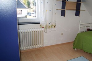 A bedroom with a radiator, a bed with a green cover, a window with curtains, and a wall-mounted shelf. - Holiday apartment with 2 bedrooms (Hooksiel \/ Wangerland)