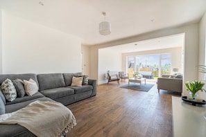A spacious living room with a grey sofa, wooden flooring, and a view through large windows. - Lantern House | Historic Wells | Sleeps 11 (Somerset)